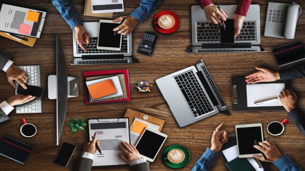 Visão aérea de uma imagem computadorizada que mostra parte dos braços e mãos de pessoas em uma mesa de reunião repleta de computadores, cadernos e xícaras de café. 