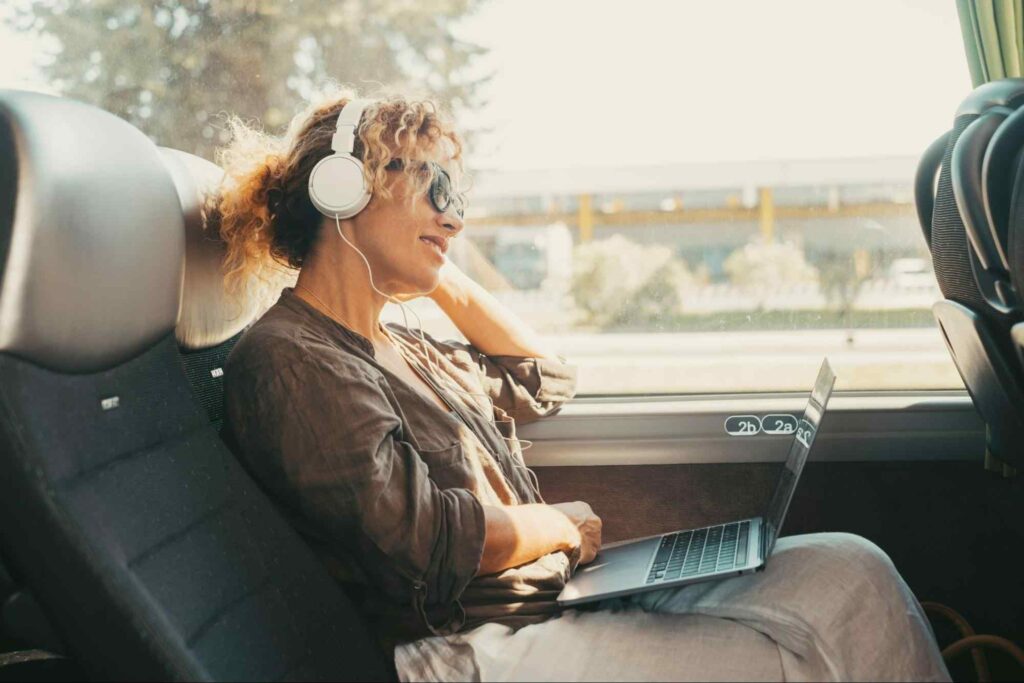 Foto de mulher sentada em uma poltrona de ônibus de viagem, com notebook no colo, headphones e óculos de sol.