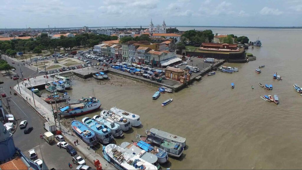 Vista aérea da parte histórica de Belém do Pará. A paisagem urbana é composta principalmente por construções antigas, como sobrados portugueses. Há uma grande praça e uma avenida larga. A cidade é abraçada pelas águas barrentas do rio, que tem várias embarcações