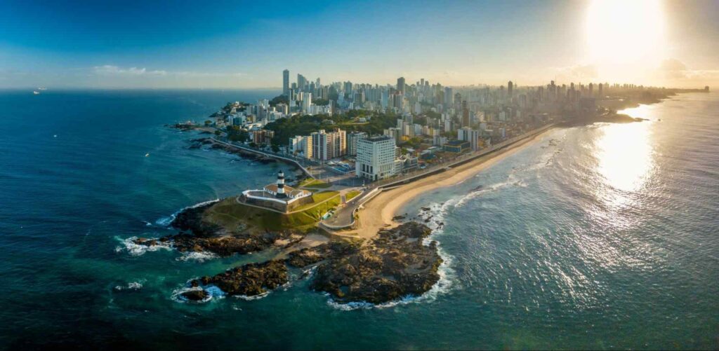 Vista aérea com a Praia e Farol da Barra (Salvador - BA) centralizados, com a cidade repleta de prédios e estabelecimentos ao fundo, cercado pelo mar azul em ambos os lados.