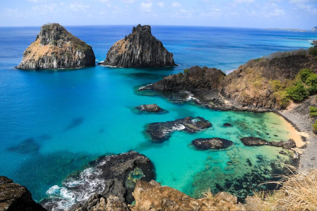 Praia da Baía dos Porcos, em Fernando de Noronha, Pernambuco. A faixa de areia é quase inexistente e a paisagem é composta majoritariamente por grandes formações rochosas em formato de monte, que são rodeadas por um mar de tom azul vibrante.