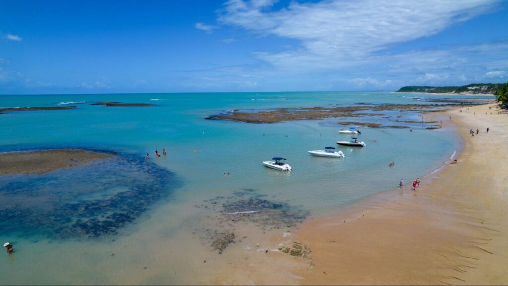 Vista panorâmica de uma das praias da Bahia. Mar azul cristalino com corais à vista, barcos na água e algumas pessoas caminhando na areia.