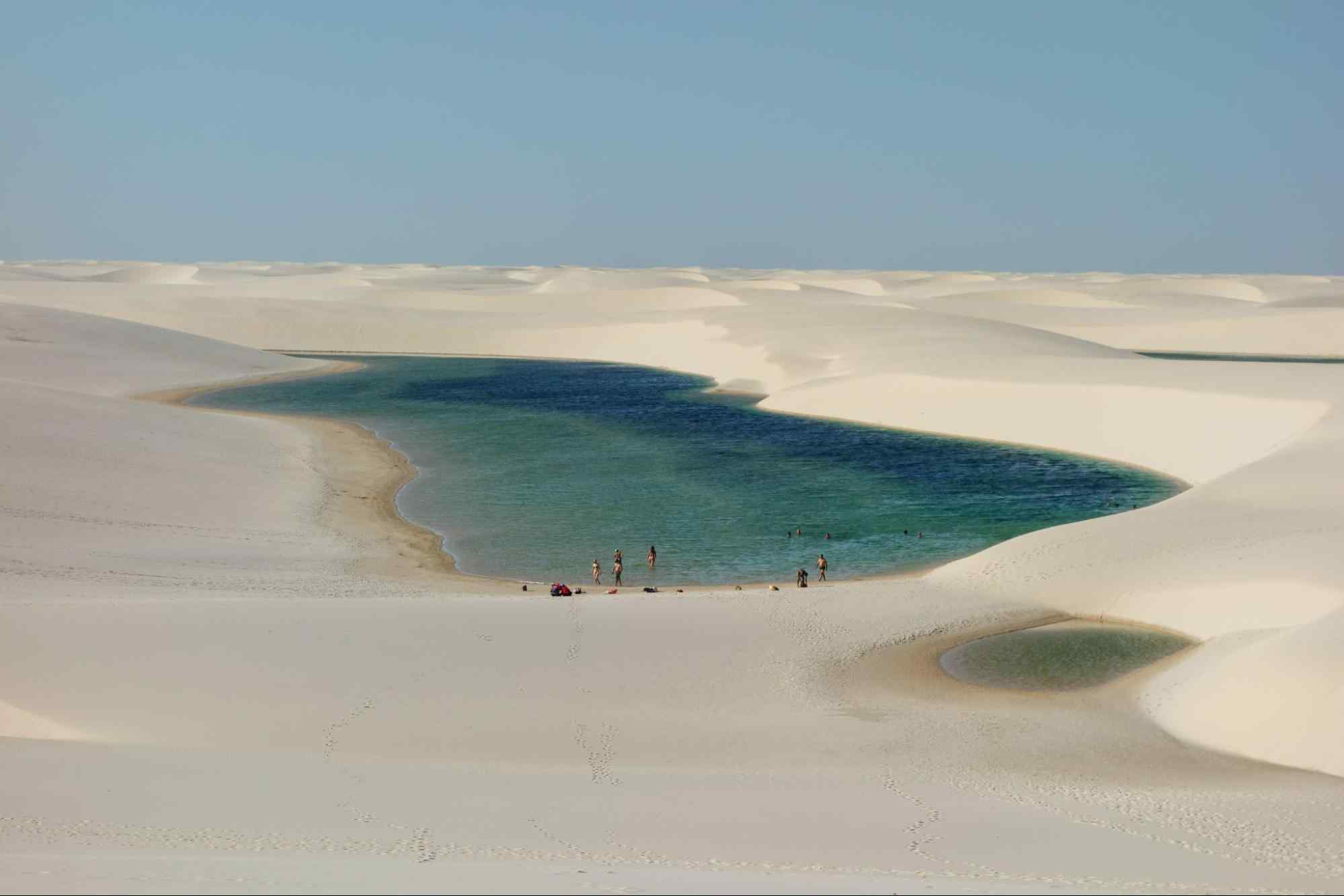 Vista aérea de uma das grandes lagoas que se formam nos Lençóis Maranhenses, dunas de areia clara ao redor da água em tons de verde e azul.