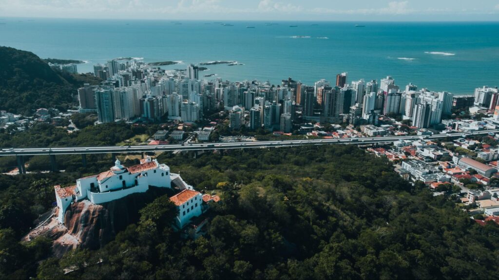 Vista aérea e panorâmica da cidade de Vitória ES. Ao lado esquerdo, Convento da Penha no topo de uma região montanhosa coberta por vegetação natural, em frente aos prédios com o oceano azul ao fundo.