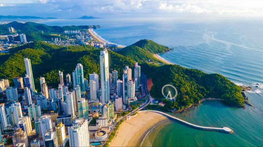 Vista aérea de Balneário Camboriú, SC. Os arranha-céus e outras grandes construções verticais dividem espaço com morros verdes cobertos de vegetação nativa. O mar que abraça a cidade é de cor azul intensa