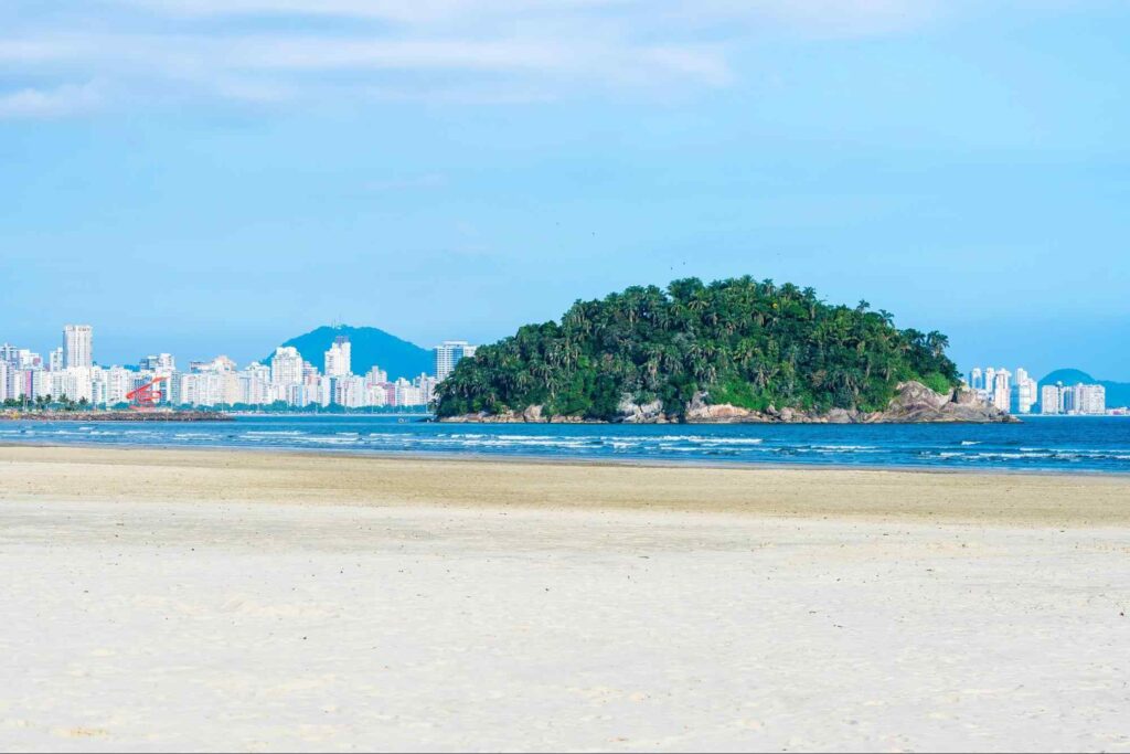 Vista panorâmica da Praia do Itararé, em Santos SP. Na imagem, céu azul, areia clara e pequena ilha repleta de árvores com prédios residenciais ao fundo da paisagem,