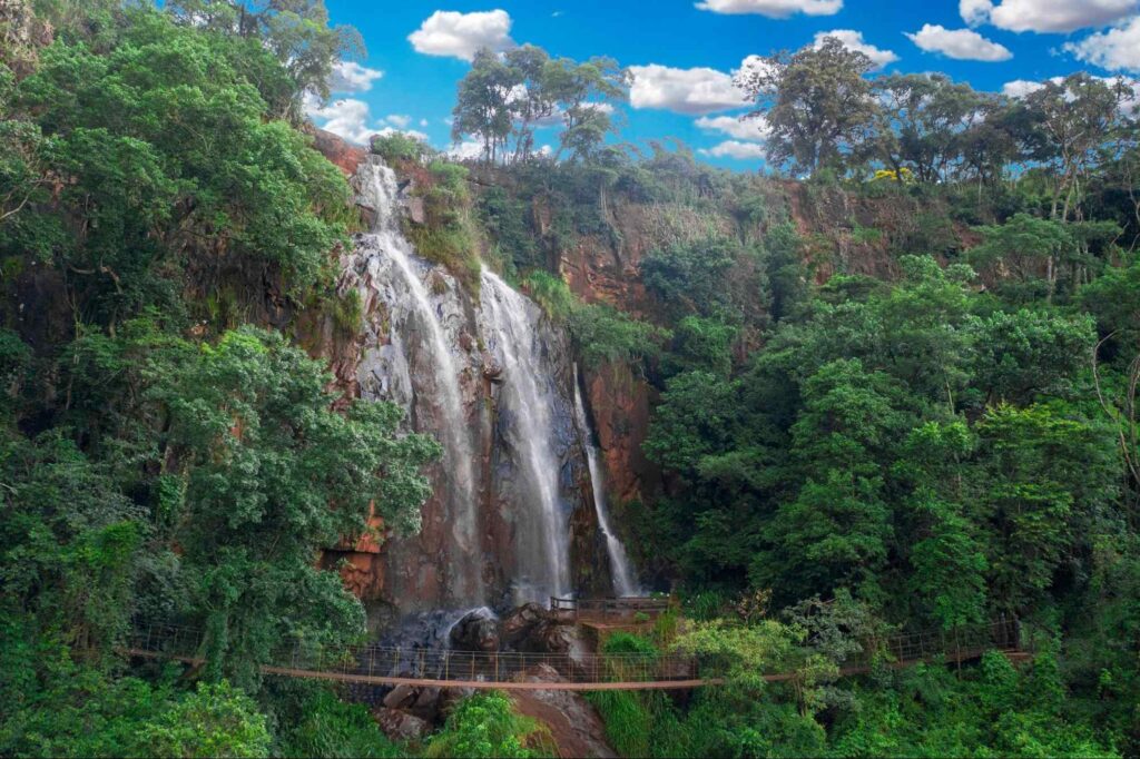 Queda d’água cercada de árvores e mata nativa, com ponte de madeira para apreciar a paisagem da queda. Céu azul com algumas nuvens brancas, em Brotas SP