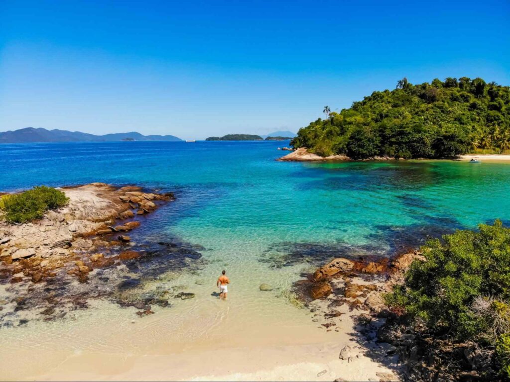 Homem entrando no mar de uma das praias de Angra dos Reis. A água é de tonalidade azul turquesa e tem algumas ilhas cobertas por vegetação verde.
