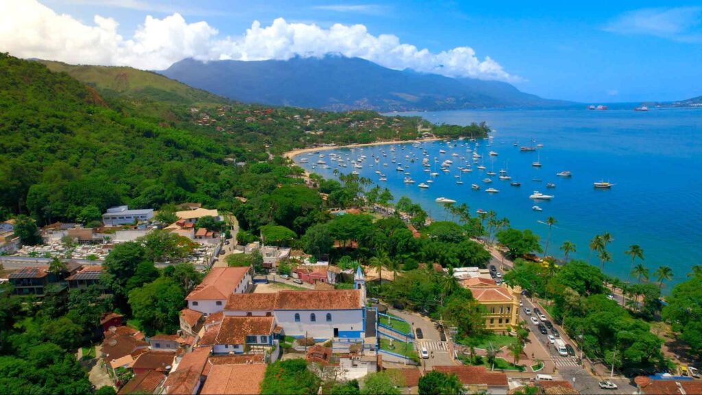 Vista aérea de Ilhabela, SP. O centro do município é composto principalmente por construções antigas e é banhado pelo mar azul. As construções estão rodeadas por árvores verdes. Ao fundo, aparecem as montanhas cobertas por vegetação nativa