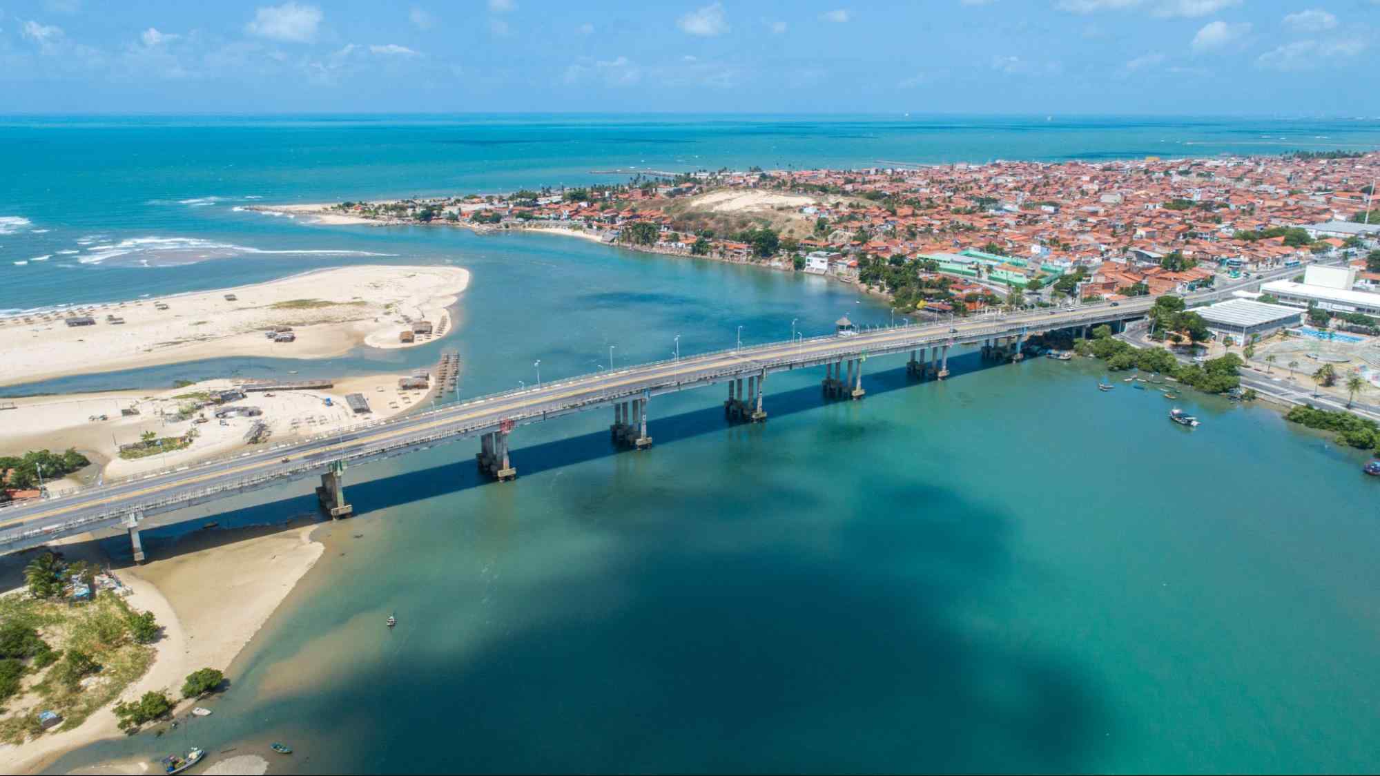 Ponte José Martins Rodrigues passando por cima das águas azuis do Rio Ceará. Ela termina na faixa litorânea de Fortaleza, CE, que aparece ao fundo.