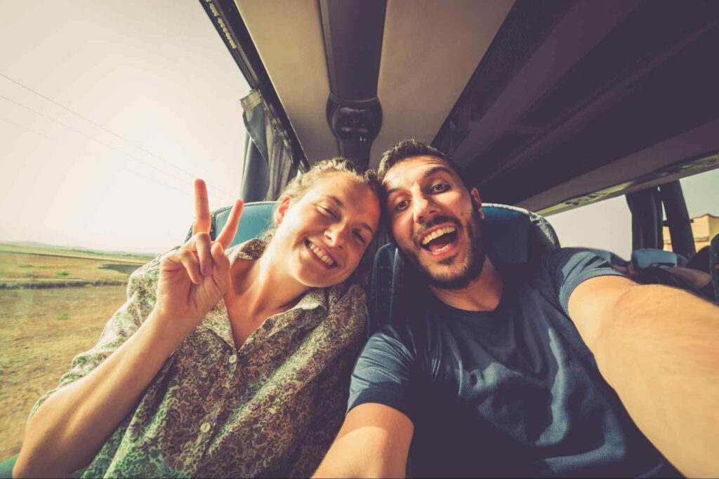 Homem e mulher sentados em poltronas de um ônibus de turismo, tirando uma selfie. A visão é da câmera, mostrando o casal e parte da paisagem pela janela.