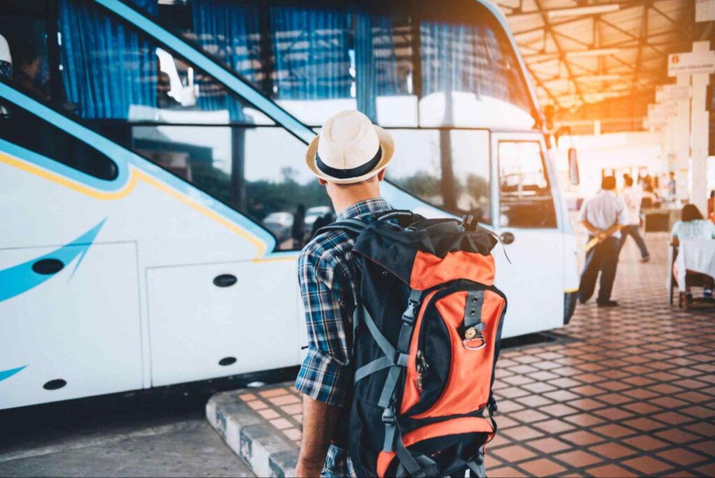 Homem de costas com uma mochila nas costas, andando em direção a um ônibus de turismo, parado em uma estação rodoviária.