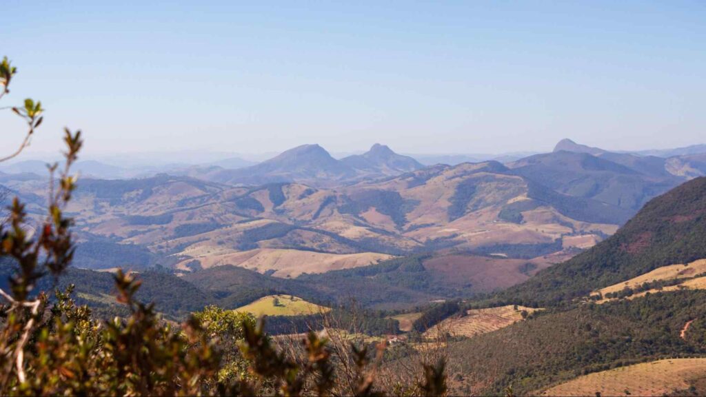 Serra da Mantiqueira e sua cadeia de montanhas, vales e campos verdes, com um céu limpo e azul ao fundo