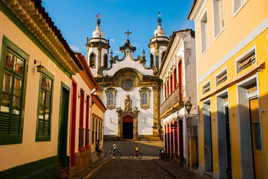 Basílica de Nossa Senhora do Pilar do ponto de vista da Rua das Casas Tortas, com chão em paralelepípedo, casas coloridas e céu azul.