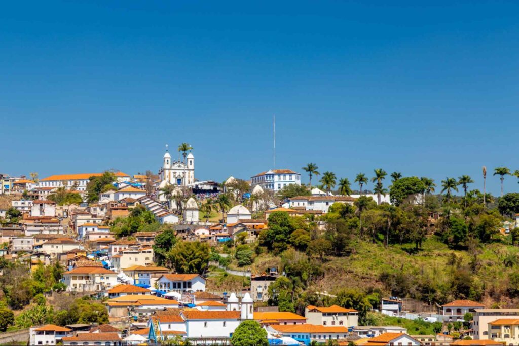 Vista panorâmica da cidade de Congonhas, Minas Gerais. Paisagem arborizada, com casarões antigos e uma igreja na parte mais alta. Ao fundo, está o céu azul sem nuvens.