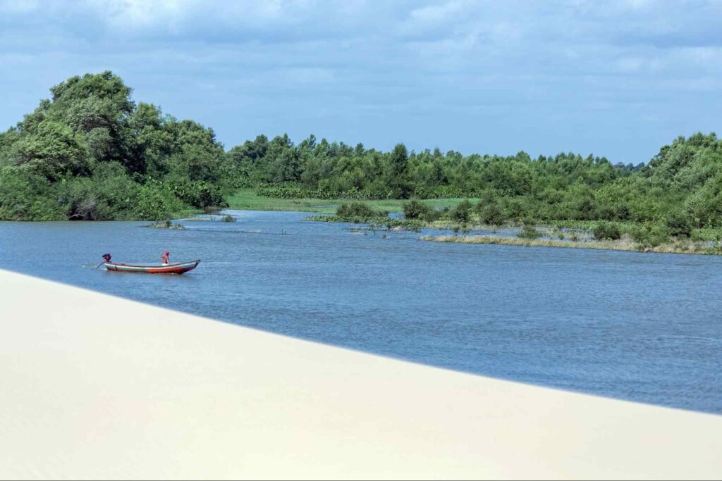 Um dos braços do Rio Parnaíba. A água é azul e contrasta com a areia branca de uma das margens e com a mata verde da outra. No canto, uma pessoa navega em um pequeno barco