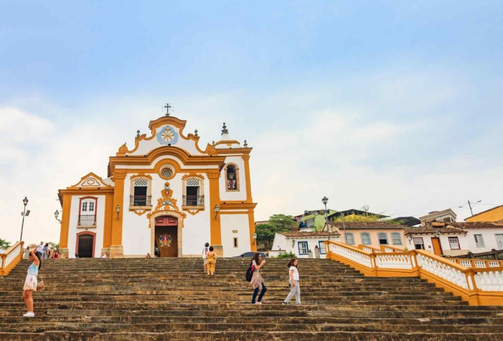 Escadaria da Igreja Nossa Senhora dos Mercês, com a igreja ao fundo. Céu limpo e azul em contraste com os detalhes amarelos da Igreja e alguns turistas em frente ao prédio.