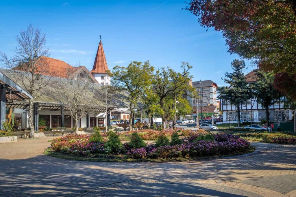 Praça em Nova Petrópolis. No centro, há um pequeno jardim circular com arbustos e flores roxas. Ao redor dele, árvores variadas e construções em estilo clássico alemão.