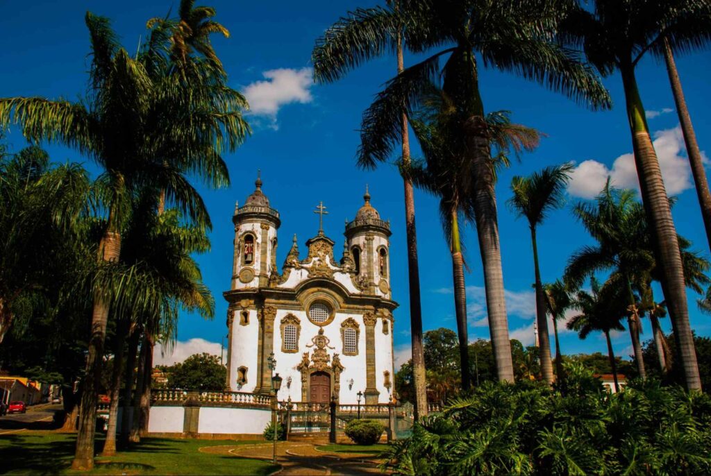 Imagem da Igreja São Francisco de Assis, dia ensolarado com céu azul, palmeiras e algumas nuvens