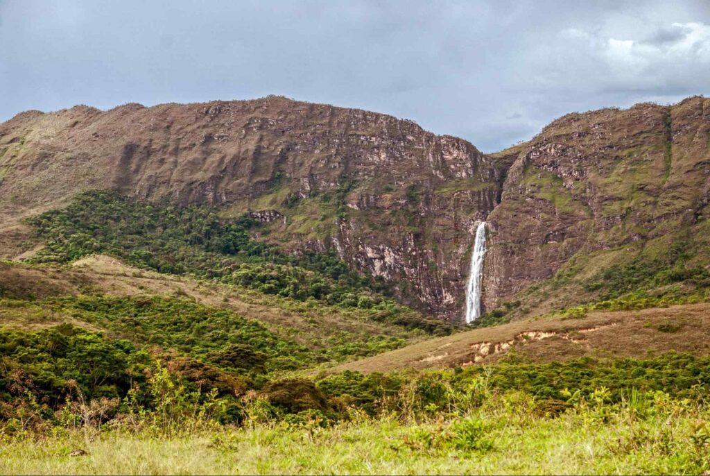 Cachoeira Casca d'Anta. Dois paredões de pedra se estendem em frente ao céu nublado. Por entre eles, uma queda d’água corre em direção a um campo de vegetação rasteira.