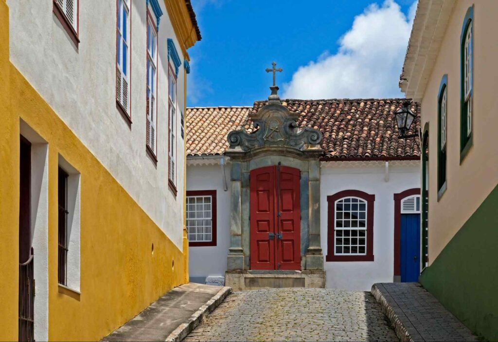 Pequena Capela de São João del Rei no final de uma rua de pedra, com portas vermelhas em destaque, paredes brancas e casas coloridas ao redor.