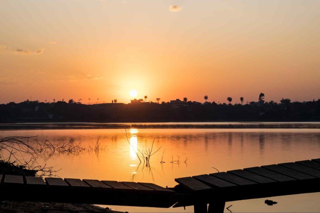 Lago em contagem com o sol se pondo no horizonte. A luz alaranjada do sol reflete na água, e uma ponte de madeira levemente quebrada aparece em primeiro plano.