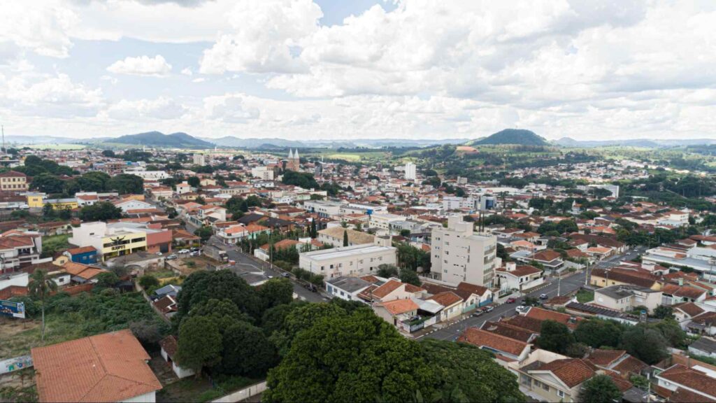 Vista aérea da paisagem urbana de Guaxupé, composta por casas e prédios baixos. As ruas são asfaltadas e bem arborizadas. Ao fundo, as serras esverdeadas e o céu encoberto por nuvens.