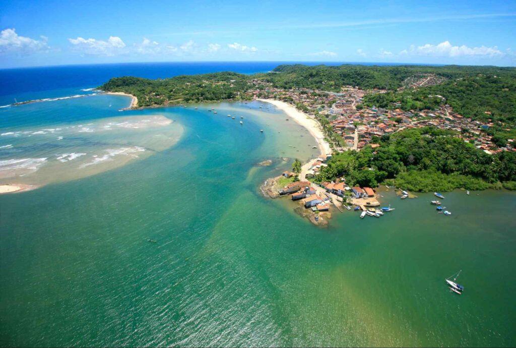 Vista aérea da linha costeira de Itacaré. No centro, faixa de areia clara se encontra com o mar esverdeado. Ao redor, casas da cidade dividem espaço com a vegetação