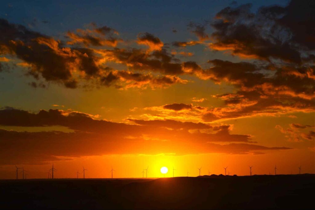 Pôr do sol no horizonte de Mossoró. O céu com nuvens se transforma de laranja para azul com a chegada da noite, e algumas torres eólicas ao fundo.