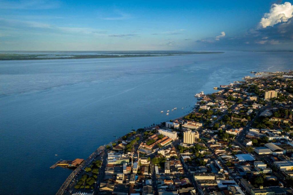 Vista aérea de Santarém. A cidade forma um contorno irregular na água azul do rio, que se estende até o horizonte. A parte terrestre exibe construções que variam entre casas e prédios baixos.