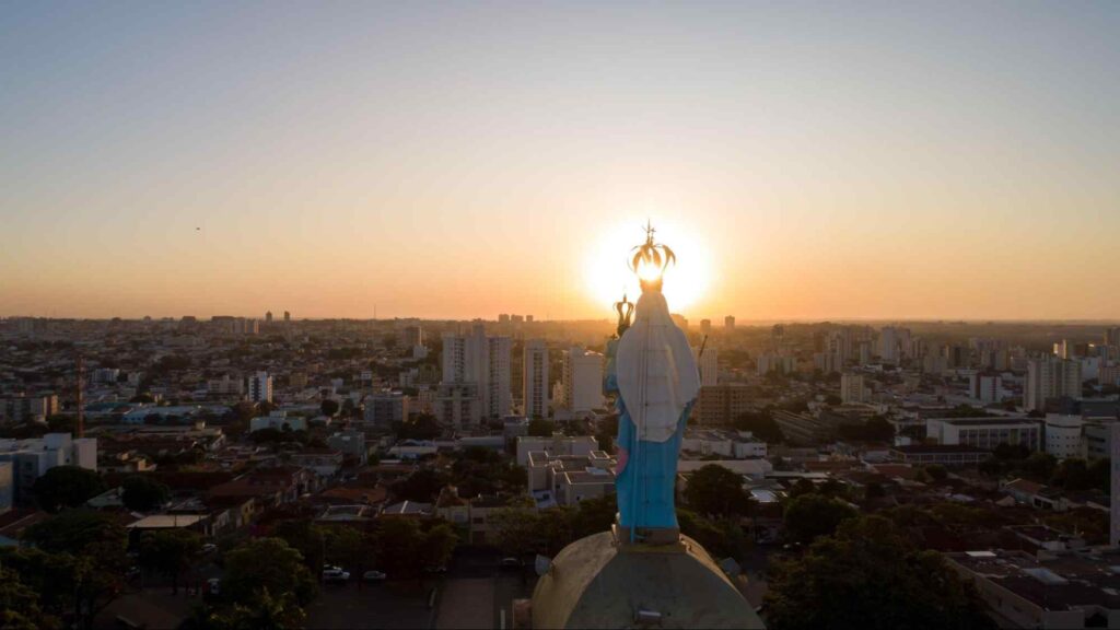 Vista aérea de Uberaba ao pôr do sol. Construções variadas e ruas arborizadas. No centro da imagem, aparece a parte de trás de uma estátua de Nossa Senhora D’Abadia.