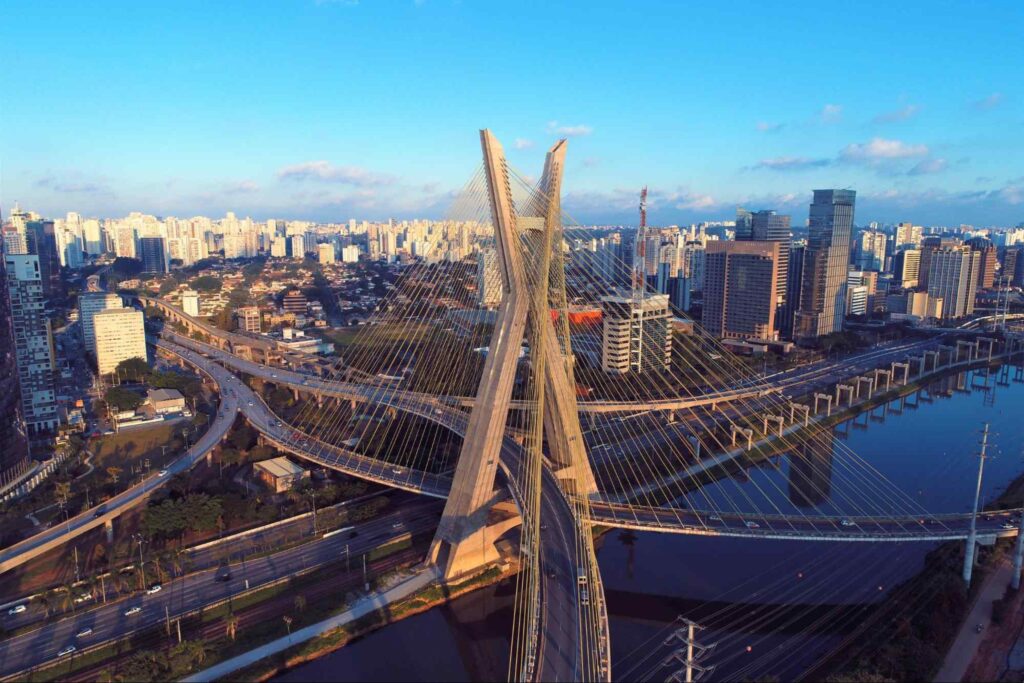 Vista aérea de São Paulo. O foco está na Ponte Estaiada, uma grande estrutura de concreto com cabos sob a qual estão pistas cheias de automóveis em movimento. Ao fundo, vê-se a paisagem urbana