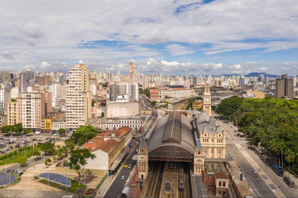 Vista aérea do bairro de Bom Retiro, em São Paulo. O destaque está na Estação da Luz, cuja estrutura abriga os trilhos do trem. Nos arredores dela, estão muitos prédios de tamanhos variados com fachadas brancas. Ao fundo, o céu azul tem muitas nuvens.