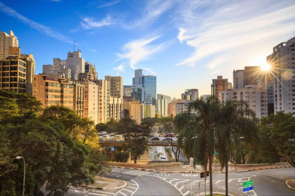 Manhã no Centro de São Paulo. As pistas repletas de carros estão rodeadas por árvores verdes. Ao fundo, vários prédios de aparência residencial. Céu está azul e tem poucas nuvens.