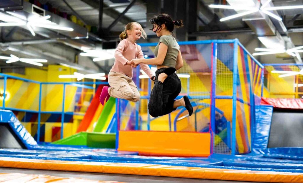 Duas garotas entre 11 e 14 anos pulando em uma grande cama elástica, de mãos dadas, uma de frente para a outra. Ao fundo, há outras camas elásticas e brinquedos coloridos.