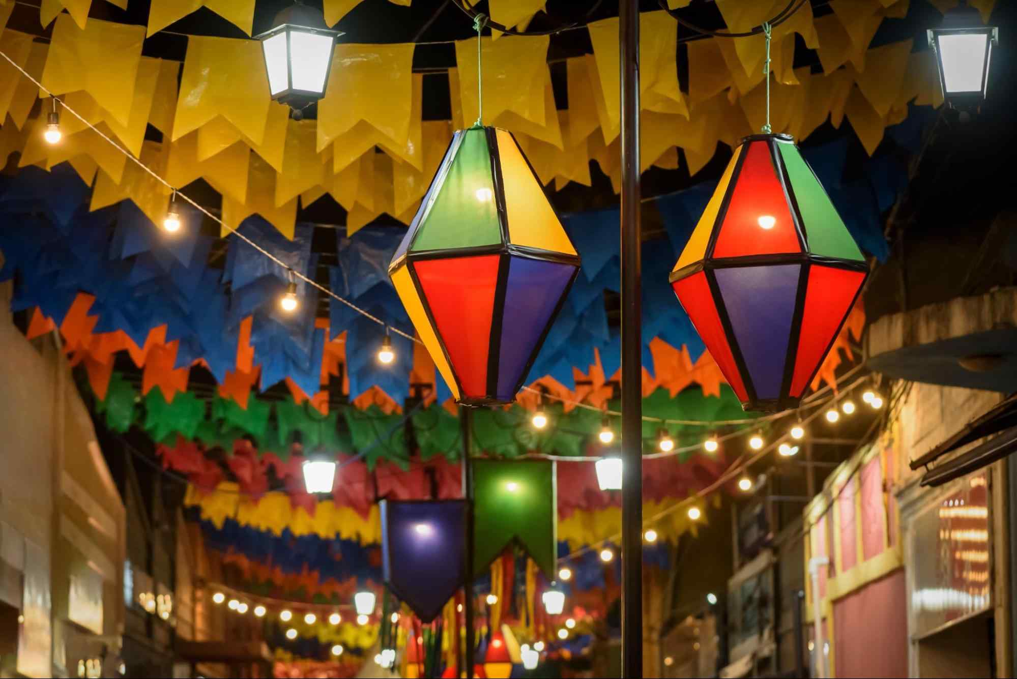 Rua decorada para o São João. Nos cordões estendidos entre as casas de um lado e de outro, estão penduradas bandeirolas coloridas, balões e luzes amarelas.