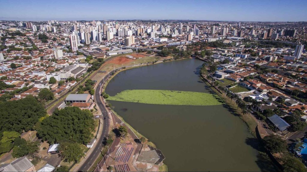 A paisagem aérea e urbana da cidade, construída ao redor de um trecho de rio com águas escuras. As construções variam entre casas, prédios baixos e edifícios de grande porte.