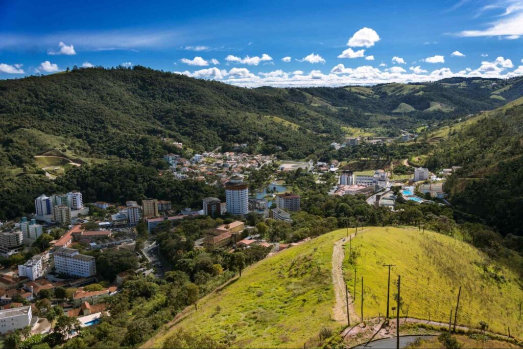 Vista aérea de Águas de Lindóia. A cidade é muito arborizada e cercada de serras verdes por todos os lados. As construções são, em sua maioria, casas e prédios baixos. Ao fundo, vê-se o céu azul e com poucas nuvens.