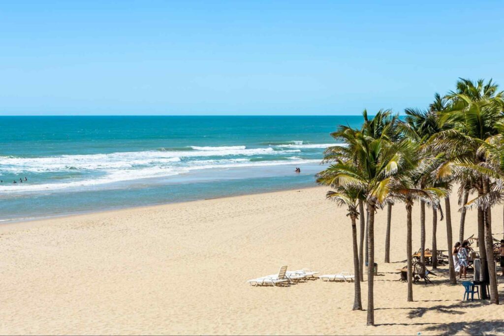 Praia de Porto das Dunas, em Aquiraz. A faixa de areia branca é larga e quase vazia, com exceção de alguns coqueiros e cadeiras de praia. O mar é azul e o céu não tem nuvens.