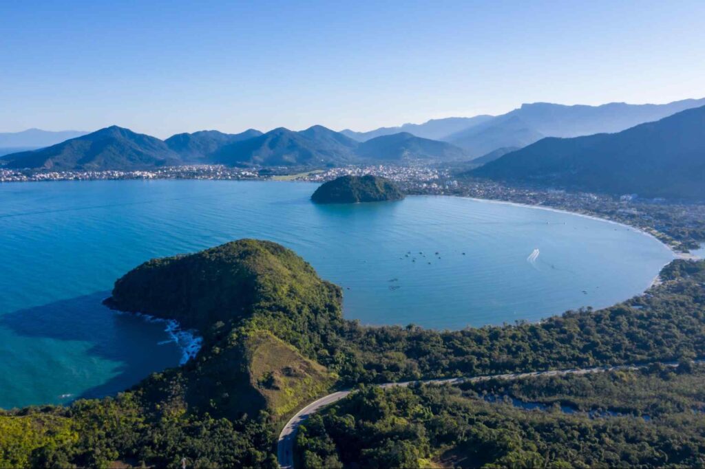 Vista aérea da costa de Caraguatatuba. O mar azul é rodeado por encostas e serras verdes e arborizadas. É possível ver as silhuetas de construções da cidade, que se aglomeram no centro da foto, ao longe.