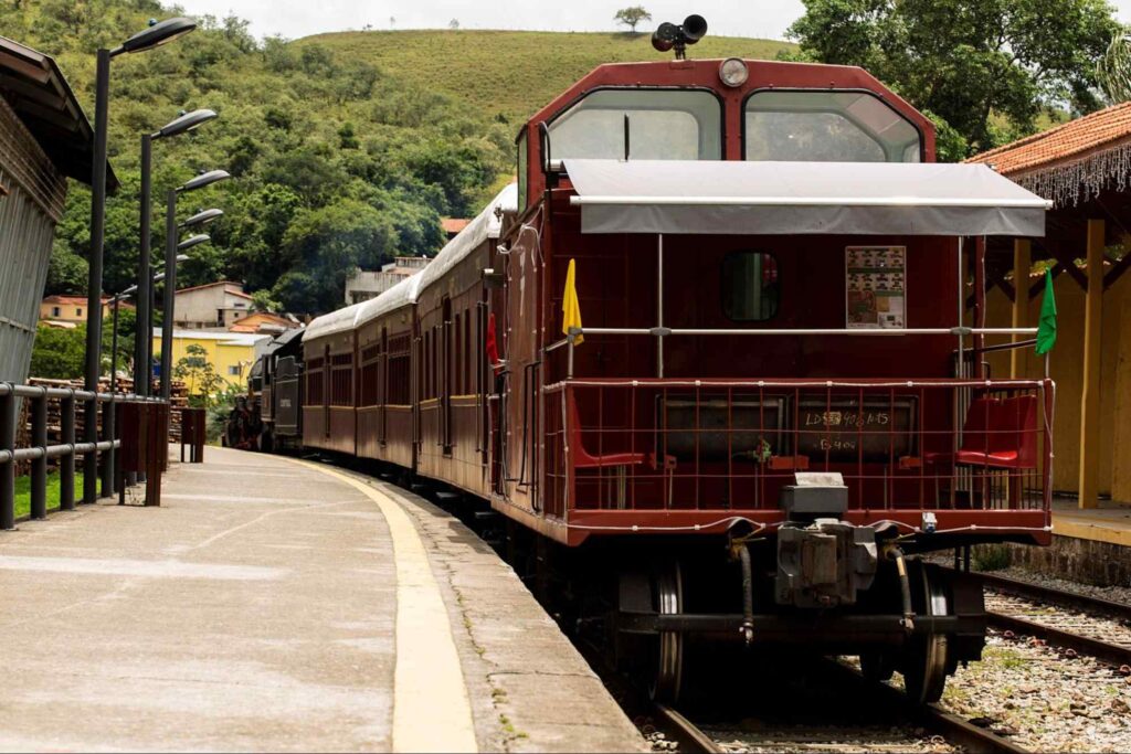 Trem vermelho parado na Estação Luís Carlos, Guararema SP. Uma passarela com postes de iluminação se destaca do lado esquerdo e ao fundo a colina com vegetação rasteira