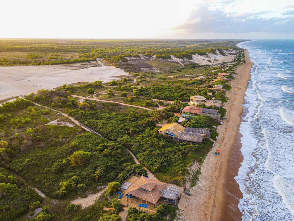 Litoral de Itaúnas, no Espírito Santo. É possível ver o mar, que chega em uma estreita faixa de areia dourada, e alguns quiosques. Depois deles, há trechos de mata verde e das dunas.