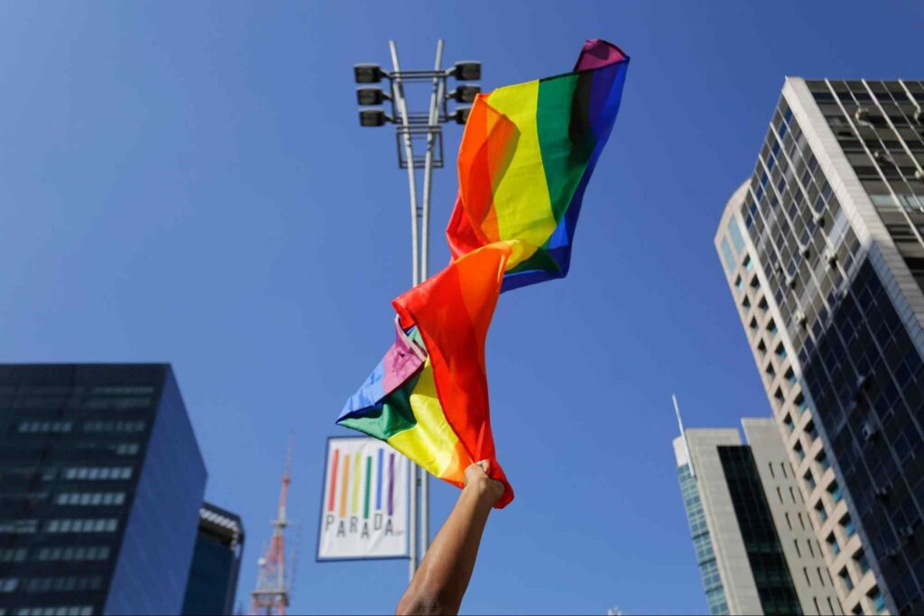 Braço de manifestante da Parada do Orgulho LGBT+ movimentando bandeira LGBT+ no ar. Céu azul com prédios ao redor da imagem.