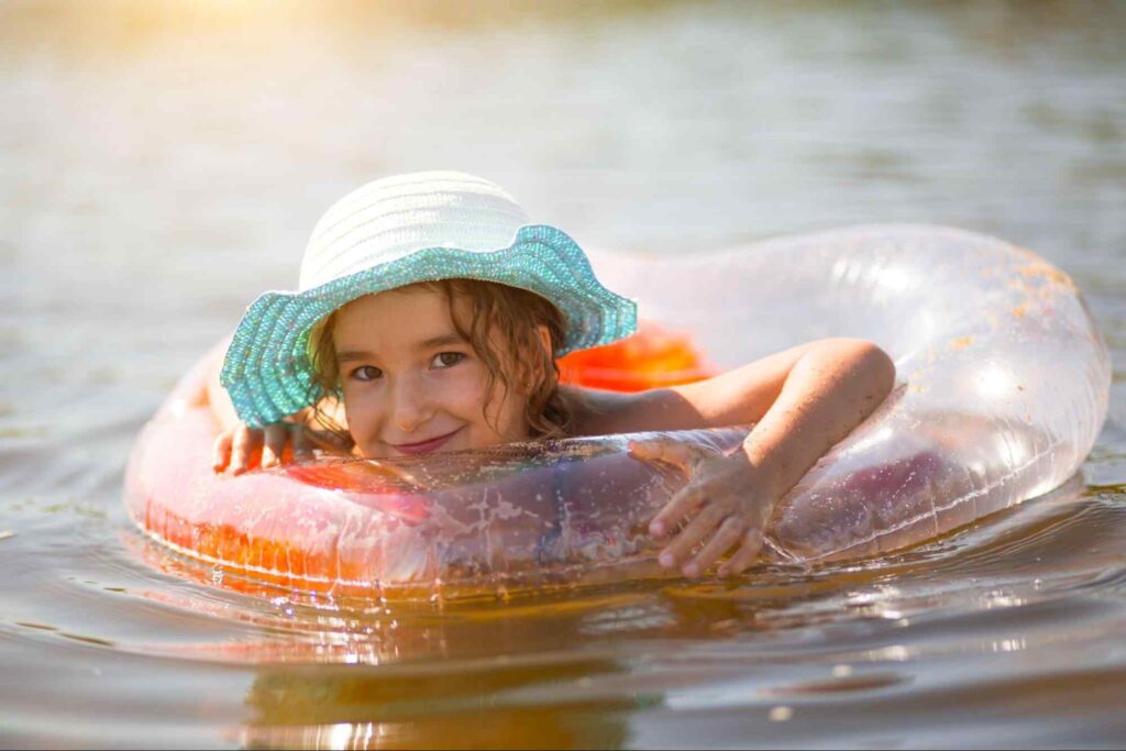 Criança em uma piscina natural, apoiada em uma boia transparente. Ela usa um chapéu e sorri.