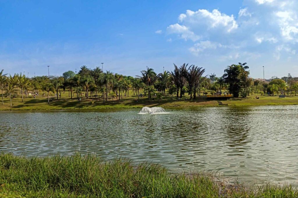 Lago do Parque Central de Santo André. Céu azul com poucas nuvens brancas em contraste com o verde das árvores e gramado ao redor do lago.