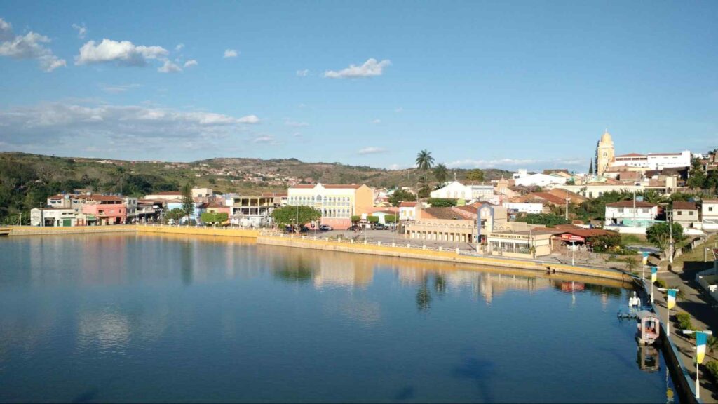 Vista aérea de Triunfo. Ao redor de um grande lago de águas escuras, estão construções baixas, como casarões e prédios de até três andares. Atrás deles, vê-se os telhados de outras casas. Ao fundo, estão as serras verdes e o céu azul, sem nuvens.