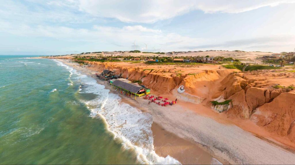 Vista aérea da Praia de Canoa Quebrada, em Aracati. O mar se encontra com uma faixa de areia. Depois dela, as falésias alaranjadas. Ao fundo, o céu está encoberto por algumas nuvens.)