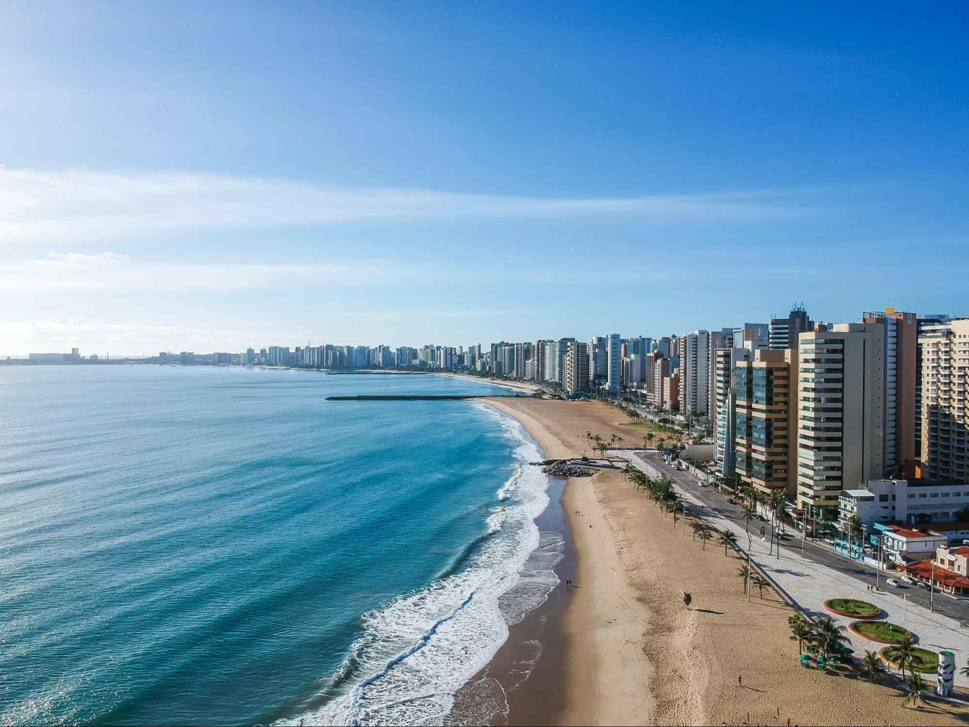 Vista aérea da Praia de Iracema, em Fortaleza. Praia urbana de mar azul e uma longa faixa de areia. A orla é delineada por prédios. Ao fundo, o céu azul aparece com poucas nuvens.