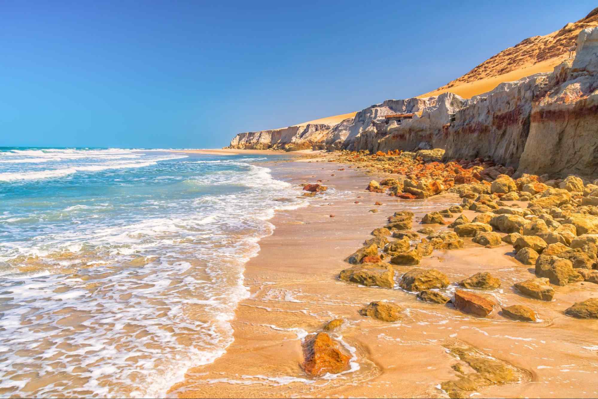 faixa de mar chegando na areia da praia de Beberibe, que está tomada por pequenas pedras do paredão de falésias que se estende no lado direito até o horizonte.