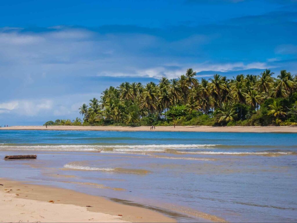 Praia do Moreré, na Ilha de Boipeba. Um banco de areia que é separado de outra faixa de areia pelas águas azuis do mar, que formam ondas discretas. Há poucas pessoas caminhando pela praia.
